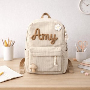 Puede incluir: Mochila de pana beige con el nombre "Amy" en letras marrones. La mochila presenta una flor blanca y un parche de seta. Tiene un bolsillo delantero y correas ajustables. La mochila está sobre una superficie de madera.