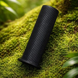 Pode incluir: Um objeto cilíndrico preto com uma textura canelada, repousando sobre uma cama de musgo verde. O objeto tem uma base alargada e um acabamento liso e escuro. O fundo apresenta vegetação exuberante, criando uma estética natural e terrosa.