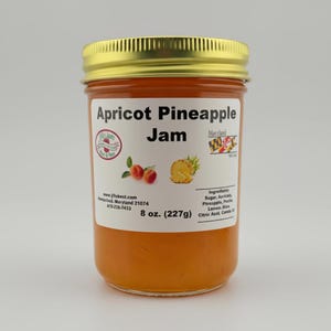 May include: A jar of apricot pineapple jam with a gold lid. The label reads "Apricot Pineapple Jam" and includes images of apricots and pineapple. The jar contains 8 oz (227g) of jam.