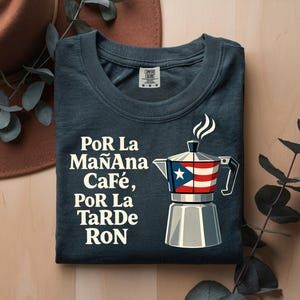 May include: A dark gray t-shirt featuring a graphic of a moka pot with a Puerto Rican flag design on its center section. The text printed in white reads: PoR La MaÑAna CaFé, PoR La TaRDe RoN. The shirt has a crew neckline and a visible brand label at the neck.