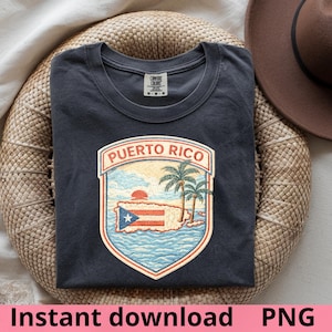 May include: Dark grey t-shirt featuring a Puerto Rico graphic. The graphic includes a shield design with the words "Puerto Rico", a map of the island, a flag, palm trees, and a sunset. The shirt is folded on a woven surface.