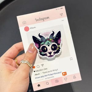 May include: A colorful sticker featuring a stylized skull with horns, stars for eyes, and floral accents. The sticker is held against a pink Instagram-themed background. The text on the background includes "Instagram" and "Album."
