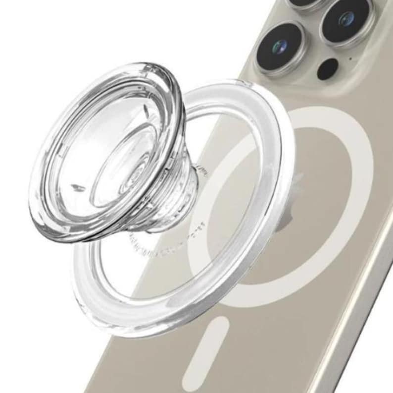 May include: A clear, circular phone grip with a transparent base, attached to a smartphone. The grip is designed to be collapsible and features a textured surface for enhanced grip. The phone case is a light beige color.