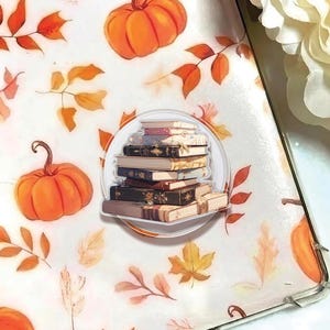 May include: A clear sticker featuring a stack of vintage books with decorative covers. The sticker is set against a white background adorned with orange pumpkins and autumn leaves. The overall theme is fall and reading.