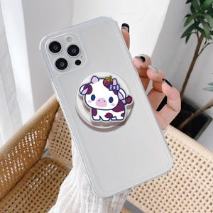 Könnte beinhalten: Transparente Handyhülle mit einem Cartoon-Kuh-Design. Die Kuh ist weiß mit lila Flecken, einer rosa Nase und einer blauen Schleife. Die Handyhülle hat einen Pop-up-Griff auf der Rückseite.