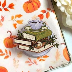 Peut inclure: Un support de téléphone en acrylique transparent avec une pile de livres et une tasse à thé. Les livres sont marron et verts avec des détails dorés. La tasse est violette avec une soucoupe verte. Le fond présente des citrouilles et des feuilles oranges.