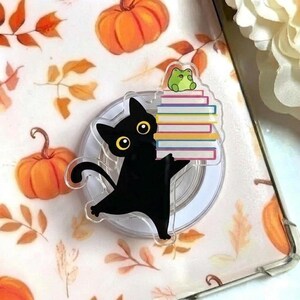 May include: A black cat-shaped phone grip with yellow eyes, holding a stack of colorful books, topped with a green frog. The grip is on a clear, circular base. The background features pumpkins and autumn leaves.