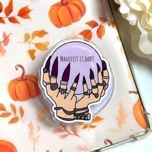 May include: A clear phone grip with a design featuring a pair of hands with long, purple fingernails holding a light purple crystal ball. The crystal ball has the text "MANIFEST IT, BABE!". The background has orange pumpkins and fall leaves.