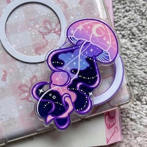May include: A clear acrylic charm featuring a stylized jellyfish design. The jellyfish has a pink, purple, and blue color scheme with a crescent moon and stars. The charm is attached to a white ring.