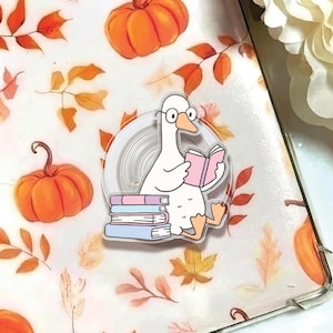 May include: A clear sticker featuring a cartoon goose wearing glasses and reading a pink book, sitting on a stack of books. The background has pumpkins and fall leaves.