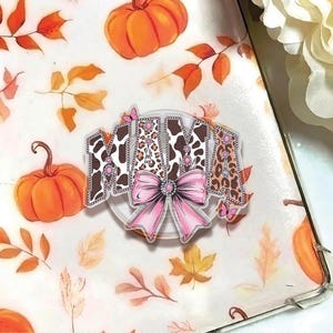 May include: A clear acrylic charm with the word "MAMA" in a mix of cow print and leopard print, accented with a pink bow and butterflies. The background features pumpkins and fall leaves.