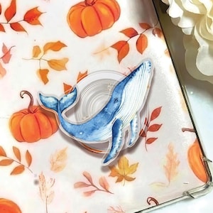 May include: A blue whale-shaped phone grip with a watercolour design. The background features pumpkins and autumn leaves in orange and yellow. The phone grip is attached to a clear phone case.