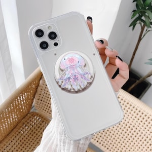 May include: Clear phone case with a decorative pop socket featuring a jellyfish design. The jellyfish is in shades of pink, purple, and white, with a flower on its head. The phone case has a clear, transparent design.