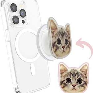 May include: A clear phone case with a magnetic ring and a cat-shaped phone grip. The grip features a close-up of a tabby cat's face with large eyes. A second cat face is shown with a pink border.