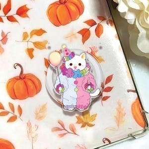 May include: A clear acrylic phone grip featuring a white cat dressed as a clown, holding a balloon. The clown costume is split pink and blue with colorful accents. The background has pumpkins and autumn leaves.
