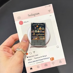 Pode incluir: Um suporte de telefone transparente com uma ilustração colorida de um armário com frascos de poções e o texto "SPACE WITHES". O suporte está preso a um cartão rosa com tema Instagram com ícones e texto de mídia social.