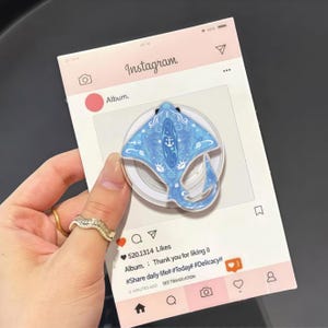 May include: A light blue stingray phone grip with a decorative design, including a white anchor, is displayed on a pink Instagram-themed background. The text on the background reads "Instagram" and "Album."