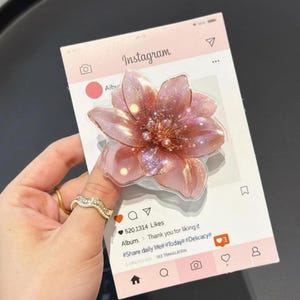 May include: A pink, glittery flower-shaped phone grip is shown on a pink Instagram-themed card. The flower has a translucent appearance with glitter accents. The card includes Instagram icons and text.