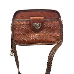 May include: Brown leather crossbody bag with a woven front panel and a silver heart-shaped clasp. The bag features a braided strap and a textured leather flap. The bag is a medium size and has a rectangular shape.