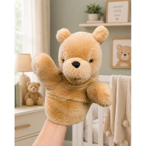 May include: A plush, tan-colored bear hand puppet with a black nose and small, round ears. The puppet's arm is raised, as if waving. The image suggests a children's toy or nursery decor.