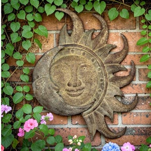 May include: A rustic metal sun and moon wall hanging with a smiling face, set against a brick wall. The sculpture has a weathered, bronze finish and is surrounded by green vines and colorful flowers.