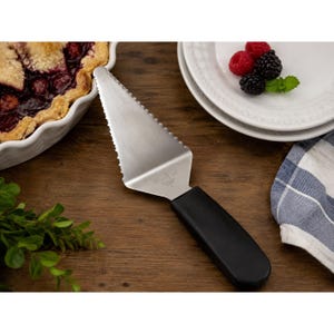May include: A stainless steel pie server with a black handle. The server has a serrated edge for easy cutting and serving. The server is on a wooden surface, with a pie and berries in the background.