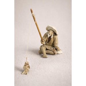 May include: A small, detailed figurine of an elderly man fishing. The man is seated, wearing a hat and traditional clothing, holding a fishing rod with a small fish at the end of the line. The figurine is in neutral tones.