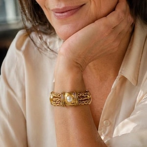 Puede incluir: Pulsera dorada con un acento de perla central. La pulsera presenta una serie de eslabones ornamentados y texturizados y una pieza central rectangular. El diseño es clásico y elegante, adecuado para ocasiones formales. Aproximadamente 18 cm de largo.