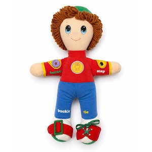 May include: A soft doll with a red top, blue trousers, and red shoes. The top has a button, snap, and a yellow circle design. The trousers have the words "buckle" and "tie". The doll has brown yarn hair and a green hat.