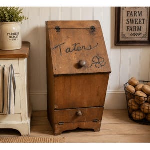 May include: A rustic wooden potato bin with a hinged lid and a drawer. The word "Taters" is handwritten on the lid, along with a carved shamrock. The bin is made of aged wood and has a dark brown finish. A basket of potatoes sits nearby.