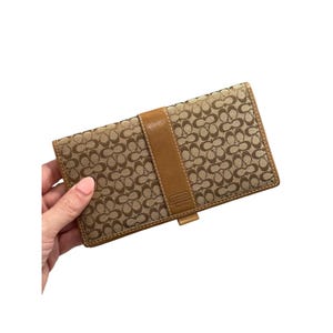 Könnte beinhalten: Beige Coach-Geldbörse mit sich wiederholendem Logomuster. Die Geldbörse hat einen hellbraunen Lederriemen und -besatz. Das Coach-Logo ist auf der Vorderseite sichtbar. Die Geldbörse wird von einer Hand gehalten.