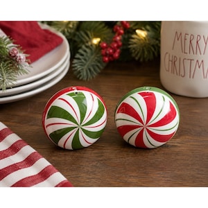 Puede incluir: Dos saleros y pimenteros redondos navideños con un diseño de remolino de menta roja, blanca y verde. Los saleros y pimenteros están sobre una superficie de madera, con una taza blanca que dice "MERRY CHRISTMAS" en el fondo.