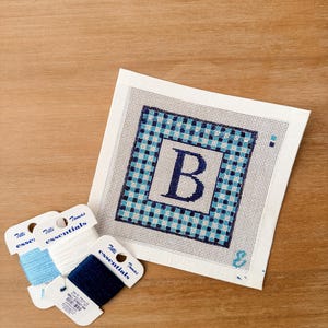 May include: A needlepoint canvas with the letter "B" in navy blue, surrounded by a blue and white checkered border. Three spools of embroidery floss in shades of blue and white are next to the canvas. The canvas is on a wooden surface.