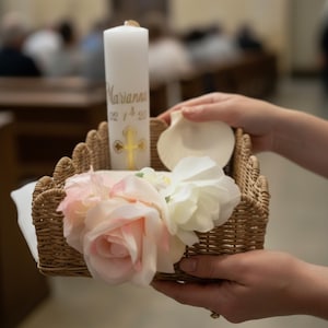 Könnte beinhalten: Eine weiße Kerze mit goldenem Text und einem Kreuz, eine Muschel und rosa und weiße Blumen in einem Weidenkorb. Die Kerze trägt den Namen "Marianna" und das Datum "02/14/20". Der Korb wird gehalten.