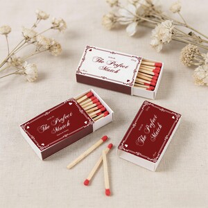 May include: Three matchboxes with the text "The Perfect Match" in a decorative font. The boxes are burgundy with white accents and contain wooden matches with red tips. Dried flowers are in the background.