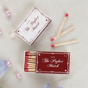 May include: A matchbox with a burgundy and white design, featuring the text "The Perfect Match" and decorative floral elements. The box is open, revealing red-tipped matches. Two loose matches are also visible.