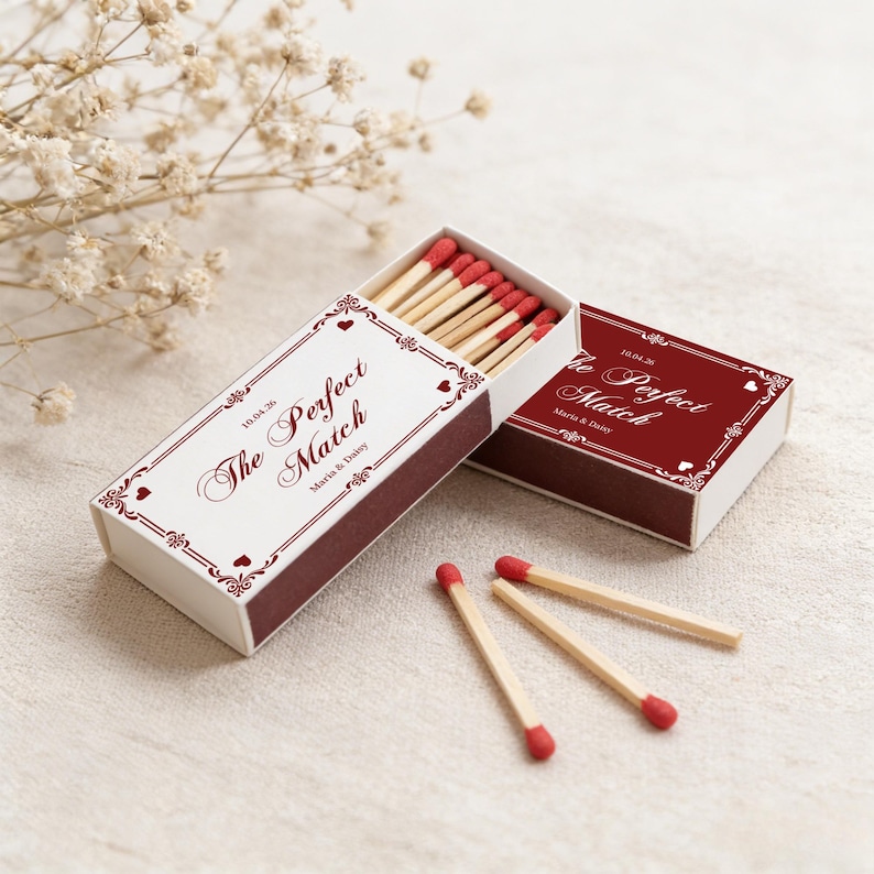 May include: Matchboxes with the text "The Perfect Match" in a decorative font. The boxes are white with burgundy trim and feature a heart design. The matches have red tips. The date "10.04.26" and names are printed on the box.