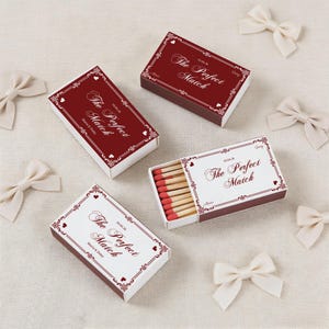 May include: Matchboxes with a burgundy and white design, featuring the text "The Perfect Match" and a date. The boxes are decorated with hearts and floral accents. Several ivory-colored bows are scattered around the matchboxes.