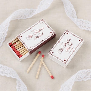 May include: Two matchboxes with the text "The Perfect Match" in burgundy script. One box is open, revealing red-tipped matches. The boxes have decorative borders and are on a neutral surface with white lace.