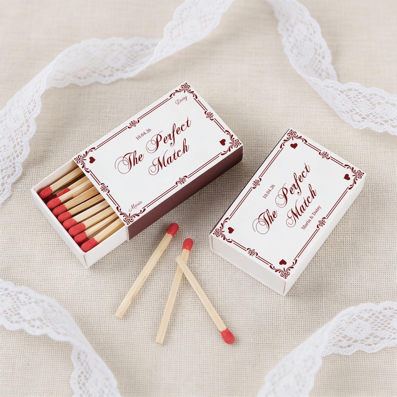 May include: Two matchboxes with the text "The Perfect Match" in burgundy script. One box is open, revealing red-tipped matches. The boxes have decorative borders and are on a neutral surface with white lace.