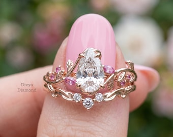 May include: A rose gold engagement ring set featuring a pear-shaped diamond and pink gemstones. The band is adorned with leaf and vine details. The ring is held up against a blurred background.