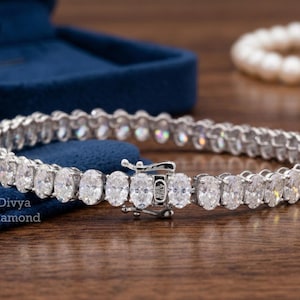 Puede incluir: Una pulsera de plata con piedras preciosas de talla ovalada. La pulsera está colocada sobre una superficie de madera, con un joyero de terciopelo azul y un collar de perlas en el fondo. La pulsera se asegura con un cierre.