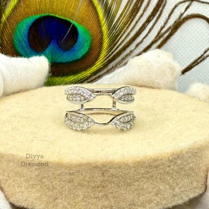 May include: A silver ring guard with a double band design, encrusted with small, sparkling gemstones. The ring is displayed on a beige, circular surface, with a peacock feather in the background. The text "Divya Diamond" is visible.