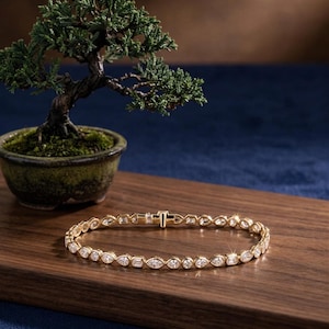 Puede incluir: Un brazalete de oro con diamantes de varias formas, incluyendo cortes de pera y baguette, se exhibe sobre una superficie de madera. Un pequeño bonsái en una maceta verde está en el fondo. El brazalete mide aproximadamente 18 cm de largo.