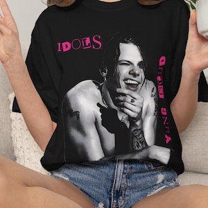 Könnte beinhalten: Schwarzes T-Shirt mit einem Schwarz-Weiß-Bild einer lächelnden Person mit Tätowierungen. Der Text "IDOLS" befindet sich über dem Bild, und "YUNG BLUD" ist vertikal auf der rechten Seite in Pink gedruckt.