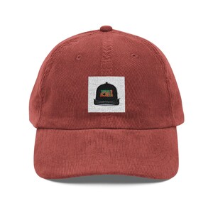 Op de afbeelding: Roodbruine corduroy baseballcap met een gebogen klep. De cap heeft een vierkante patch met een gestileerd hoedontwerp in zwart, groen en oranje. Een klassiek, casual accessoire.