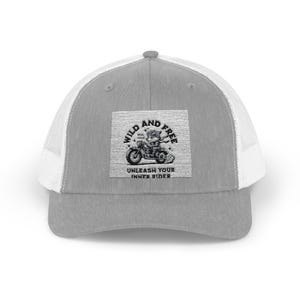 May include: Gray trucker hat with a white mesh back and a gray front panel. The front features a square patch with a black and white illustration of a bear riding a motorcycle, with the words "WILD AND FREE" and "UNLEASH YOUR INNER RIDER."
