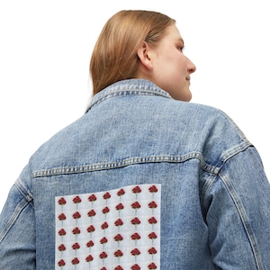 Op de afbeelding: Lichtblauwe spijkerjas met een vierkante patch op de rug met een patroon van rode rozen op een witte achtergrond. De jas heeft een klassieke kraag, een knoopsluiting en lange mouwen.