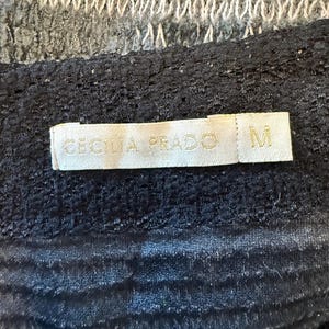 May include: A close-up of a black garment with a textured surface. A label with the text "CECILIA PRADO" and the letter "M" is visible. The top portion of the image shows a gray and white knitted fabric.