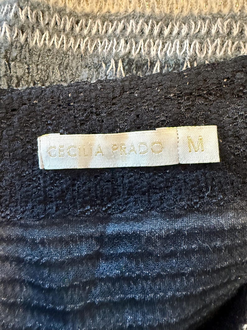 May include: A close-up of a black garment with a textured surface. A label with the text "CECILIA PRADO" and the letter "M" is visible. The top portion of the image shows a gray and white knitted fabric.
